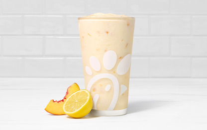 A glass of Peach Frosted Lemonade with a peach slice and a lemon half on a white countertop with a white tiled background